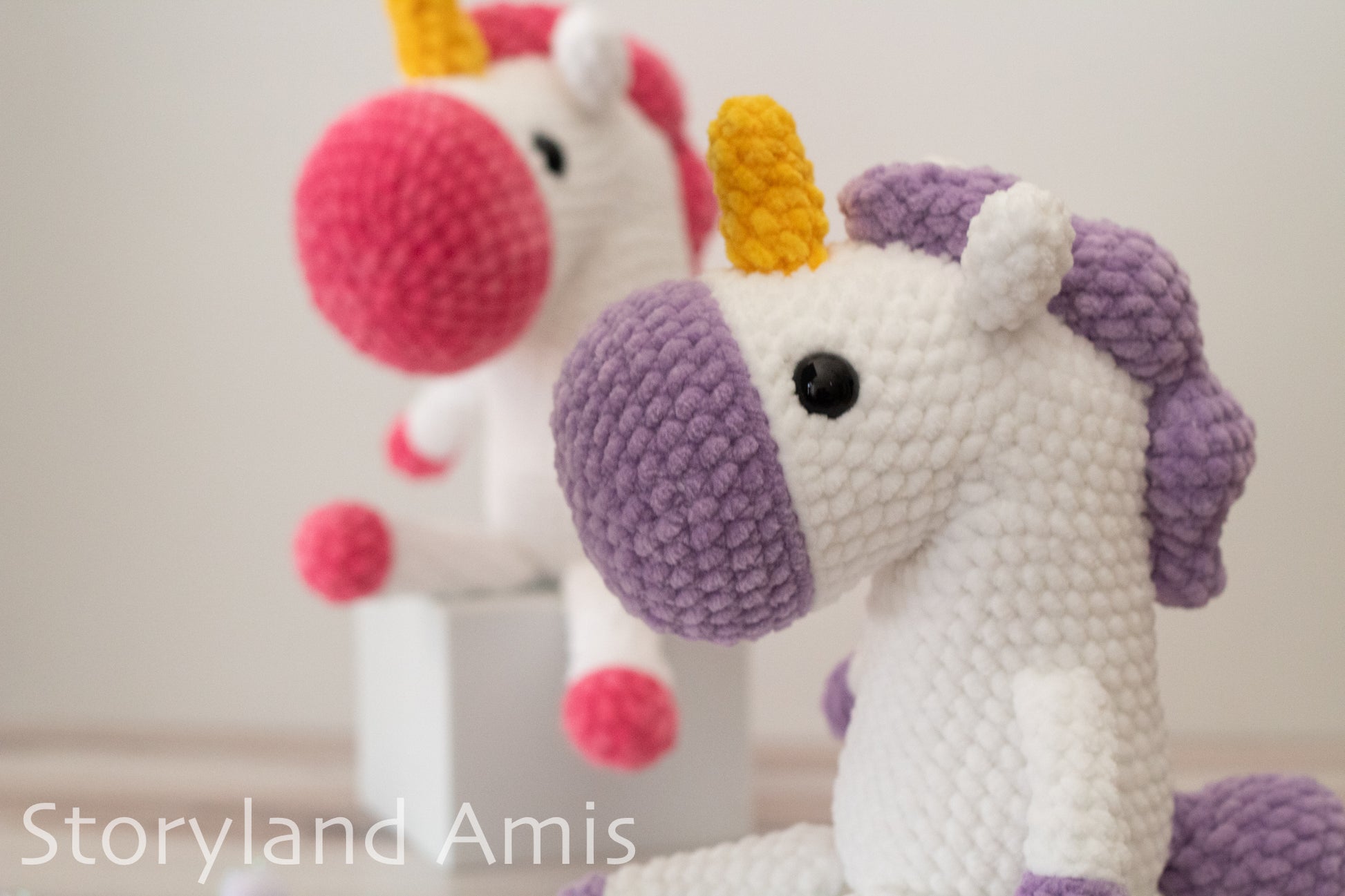 Two crochet unicorns with colorful manes on a plain background, branded 'Storyland Amis'.