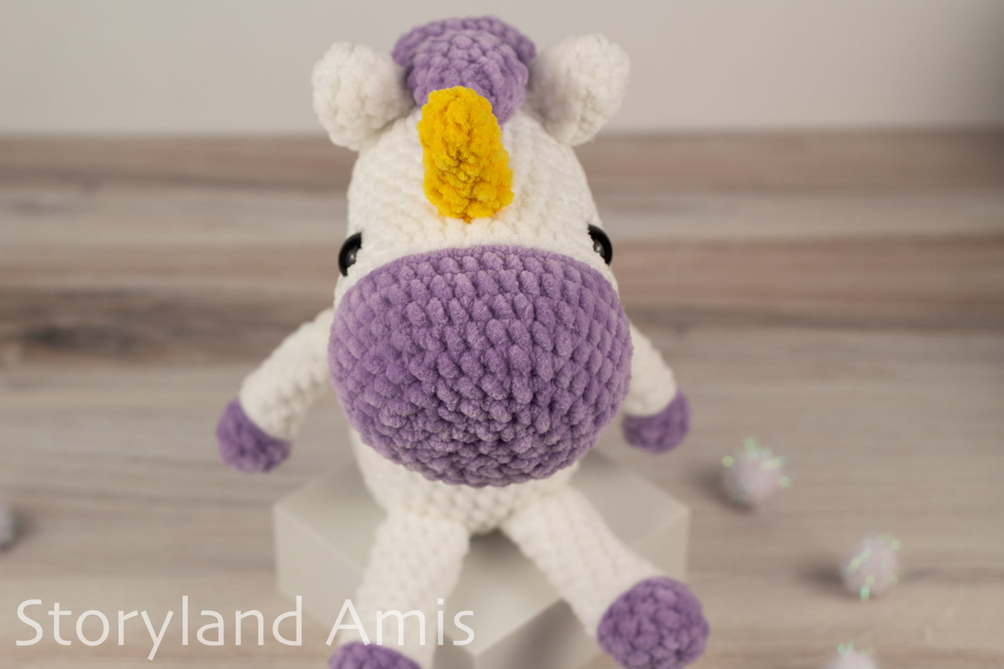 Crocheted unicorn toy with a purple horn and hooves on a wooden surface, branded 'Storyland Amis'.