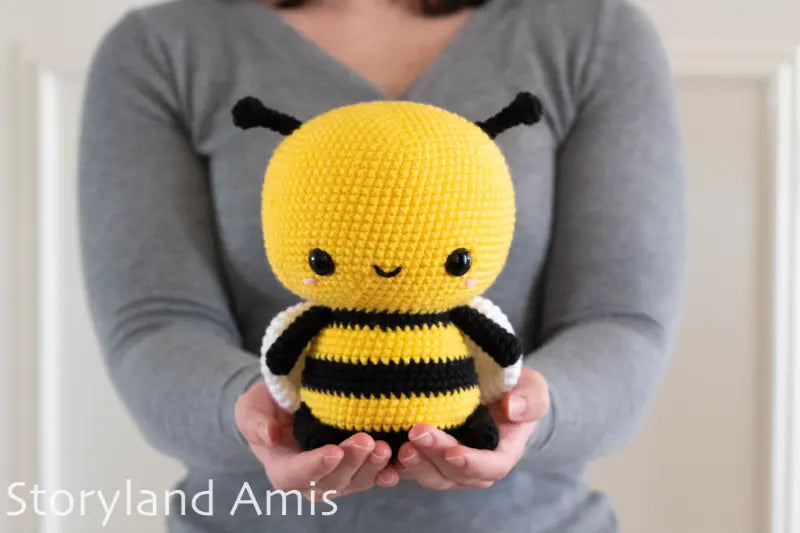 Person holding a yellow and black bee-shaped amigurumi toy with 'Storyland Amis' branding.