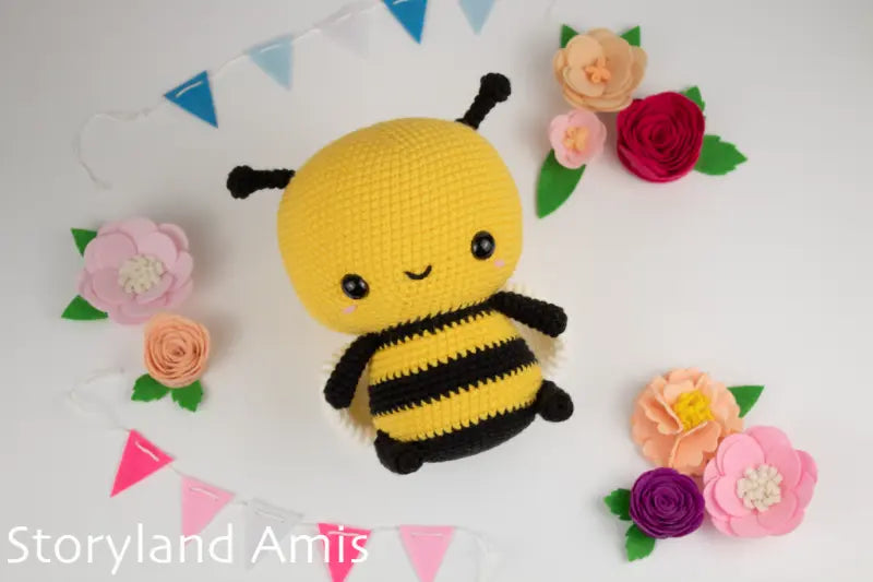 Crocheted bee toy with flowers and bunting on a white background, featuring 'Storyland Amis' branding.