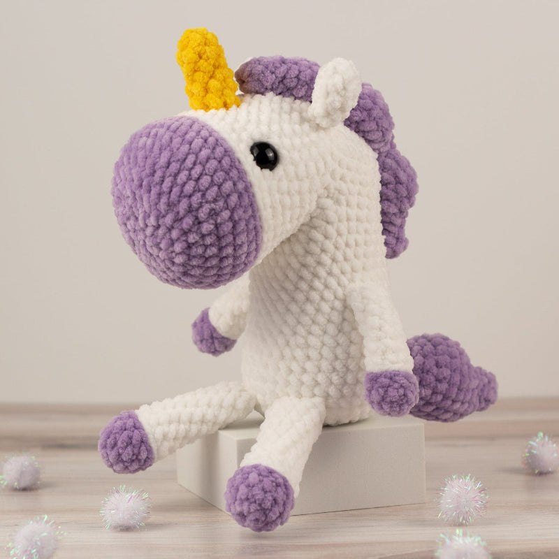 Crocheted unicorn toy with a purple body and yellow horn on a light wooden floor.