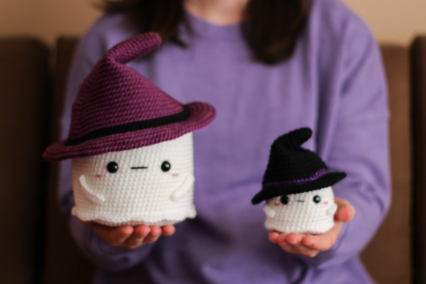 Two small crocheted ghosts with witch hats held by a person wearing a purple shirt.