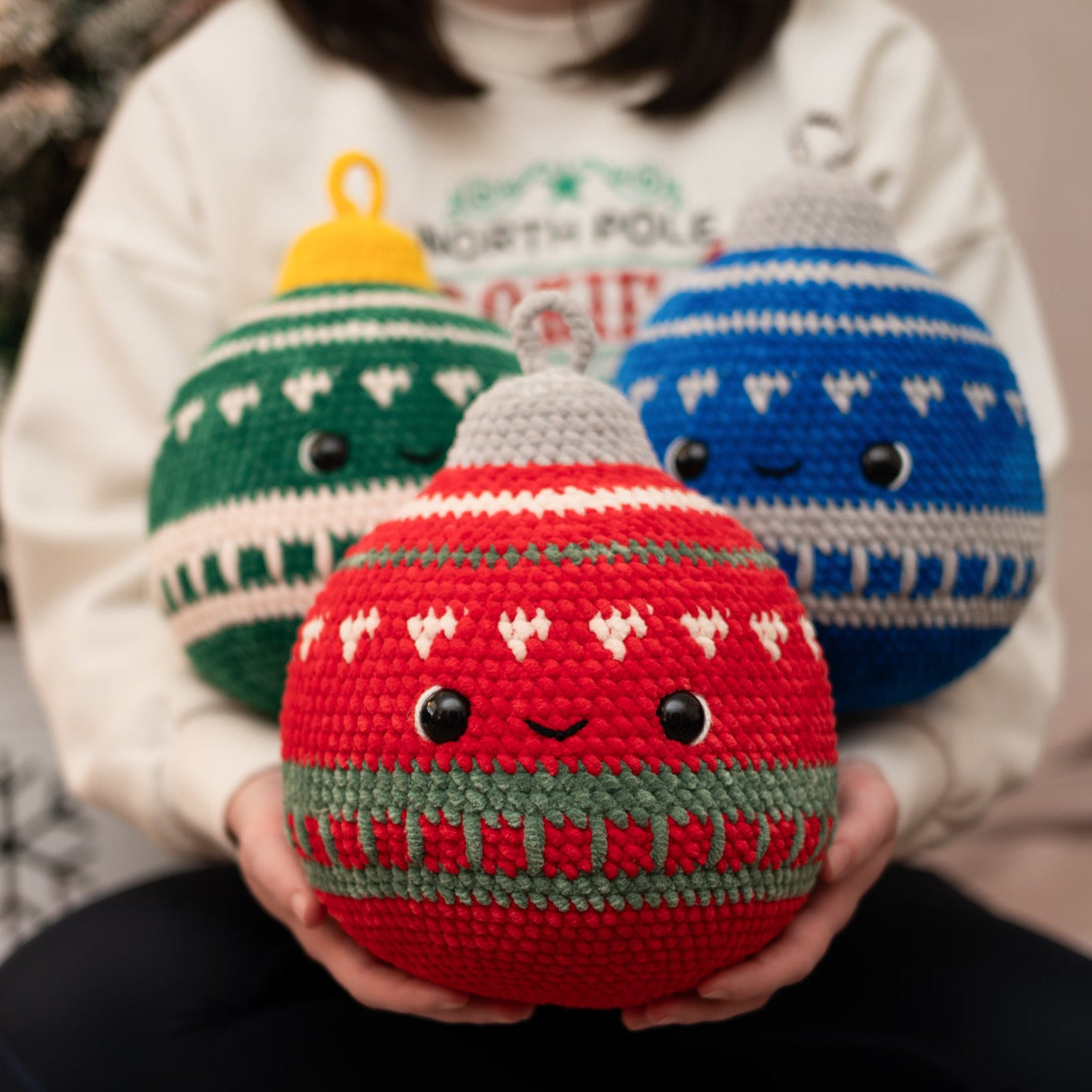 Person holding three crocheted Christmas ornaments in front of a decorated Christmas tree.