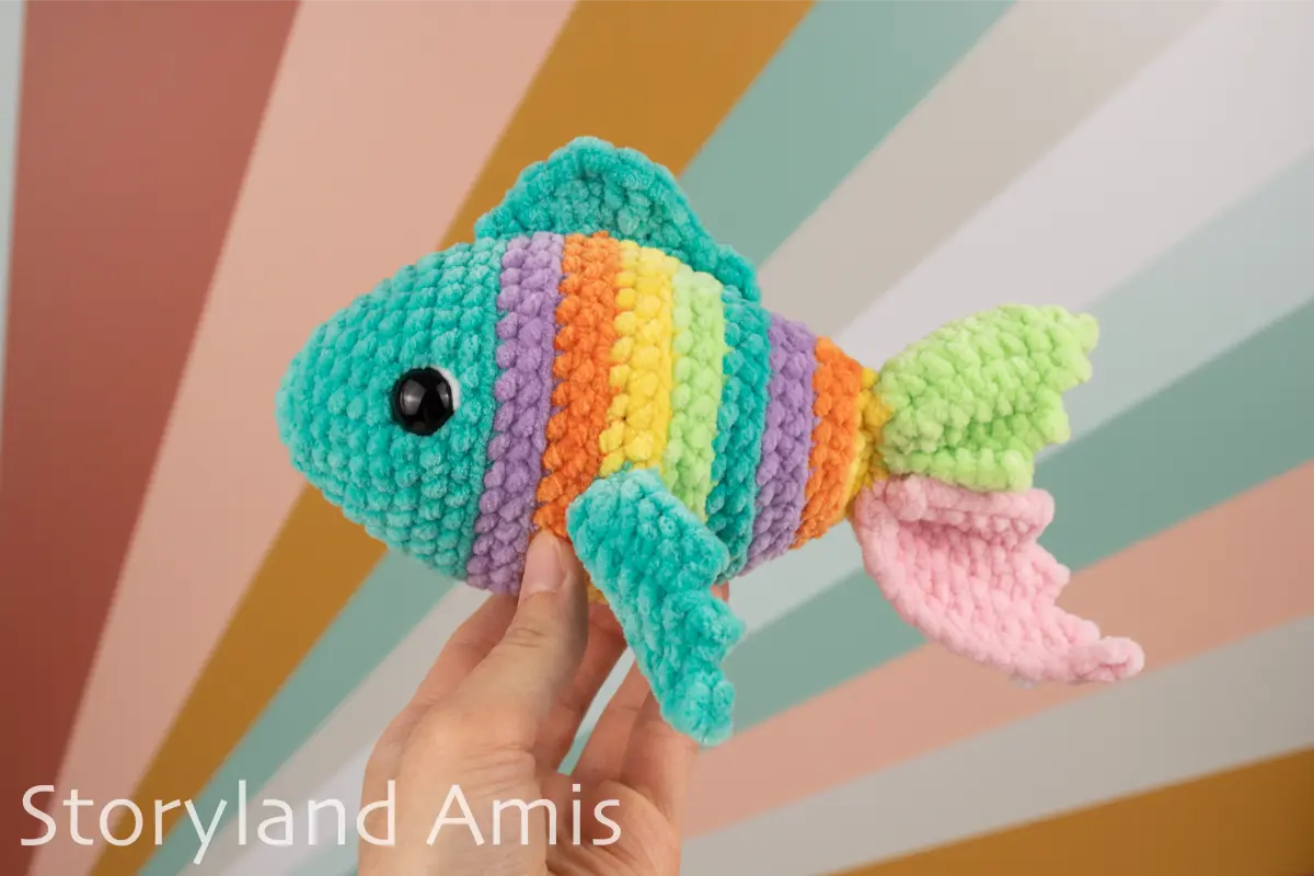 Colorful crocheted fish toy held by a hand against a multicolored striped background, with 'Storyland Amis' branding.