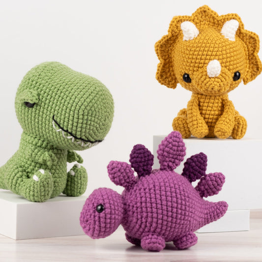 Three crocheted dinosaur toys in green, yellow, and purple on a white background